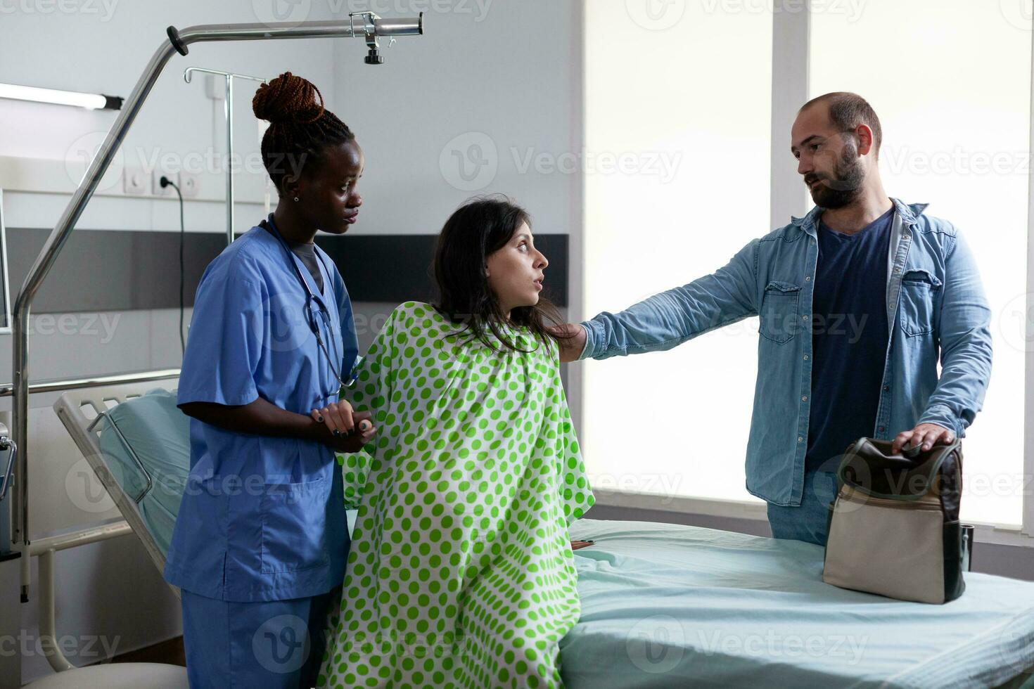 African american obstetrics nurse helping pregnant woman to get out of bed in hospital ward, preparing patient for caesarean surgery. Future parents expecting child in maternity clinic photo
