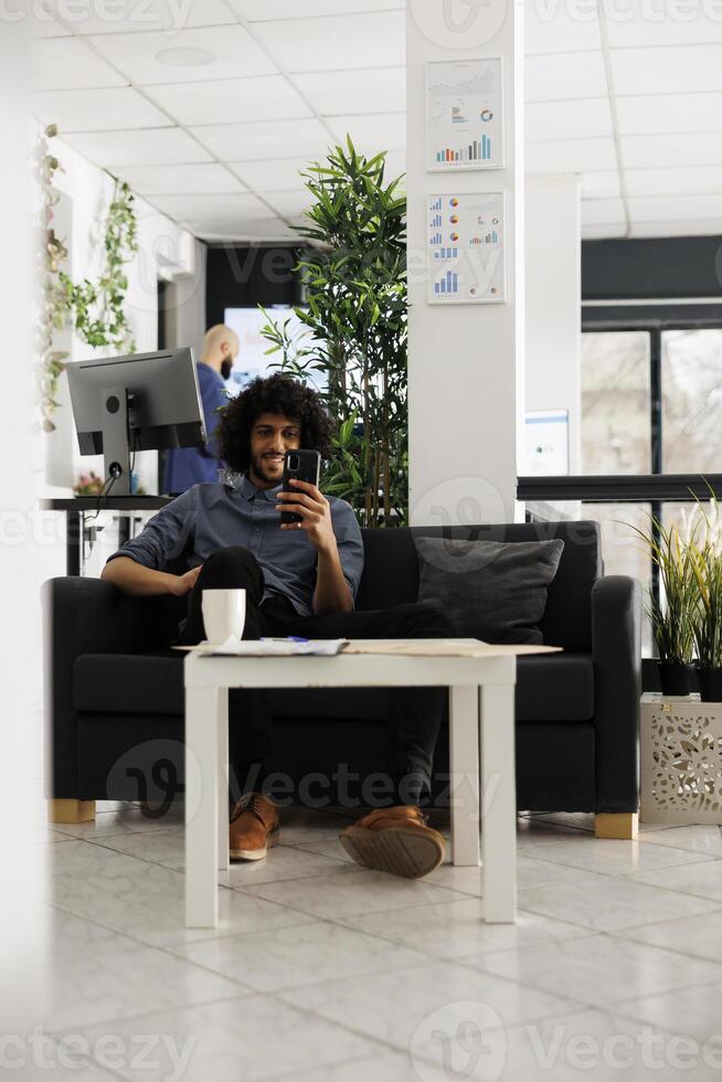 Smiling executive manager relaxing and chatting on smartphone videocall in office. Happy young arab businessman talking on video conference using mobile phone in coworking space photo
