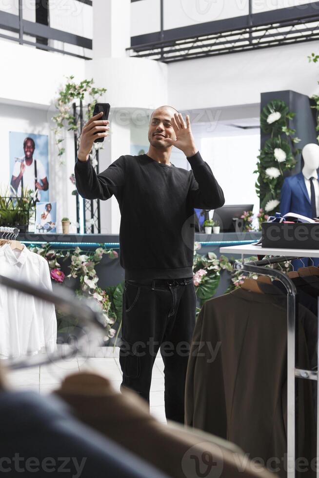 Arab man influencer promoting boutique while streaming in clothing store using smartphone. Shopping mall young customer holding mobile phone, waving hi and recording video photo