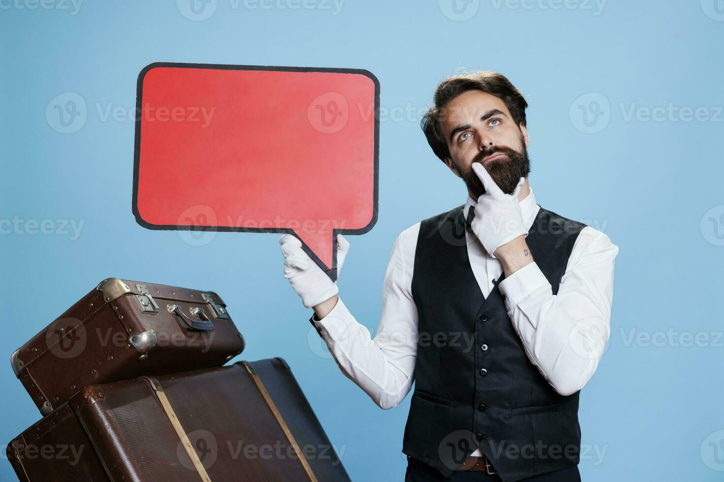 Doorkeeper holds blank speech bubble in studio, creating advertisement with isolated copyspace red cardboard icon. Bellhop with gloves and tie pointing at empty billboard sign. photo