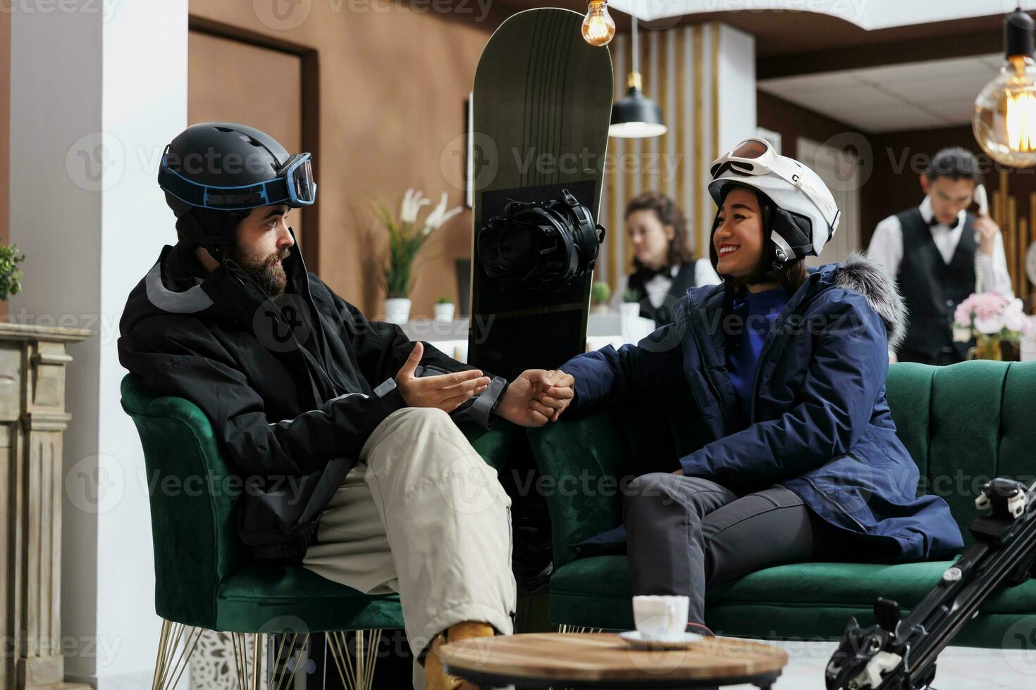 Boyfriend and girlfriend in possession of wintersports equipment engaged in dialogue about plans for skiing and snowboarding. Enthusiastic couple warmly dressed for snow activities seated in lobby. photo