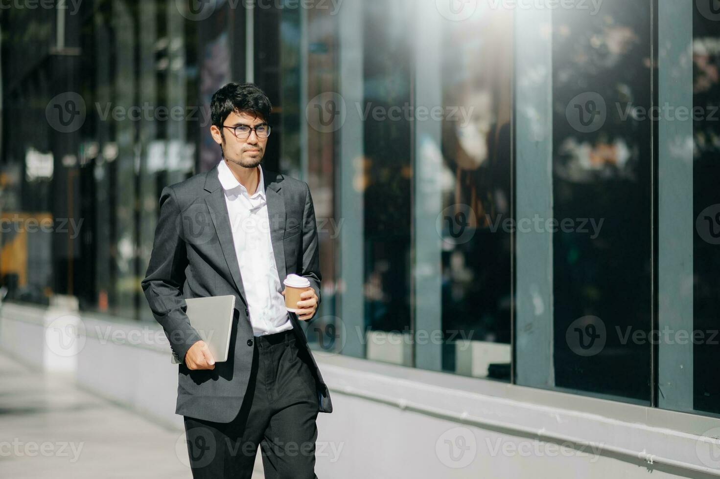 Handsome male entrepreneur standing outdoors with coffee cup and laptop in hands, leaving office in formal clothes waiting for taxi photo