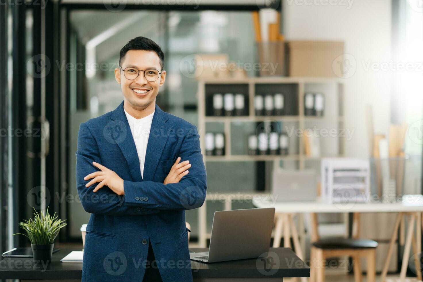 Young attractive Asian man smiling thinking planning writing in notebook, tablet and laptop working from home, looking at camera at office photo
