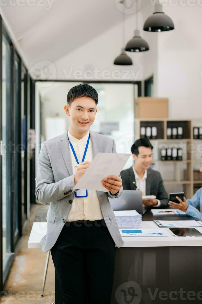 Young attractive Asian male office worker business suits smiling at camera with working notepad, tablet and laptop documents in office photo