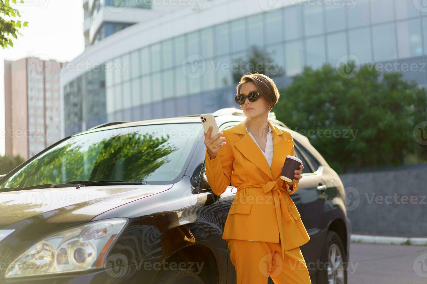 Pretty  woman with short haircut  talking mobyle phone and  posing near luxury car. Modern outdoor background.  Wearing yellow suit and sunglasses. Holding cup of coffee. photo