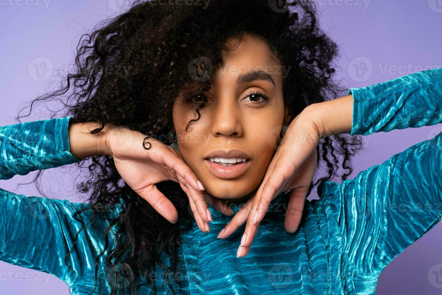 Close up  studio fashion portrait of gorgeous brazillian woman with curly hairs in elegant velvet dress over blue violet background.  hends near face. photo