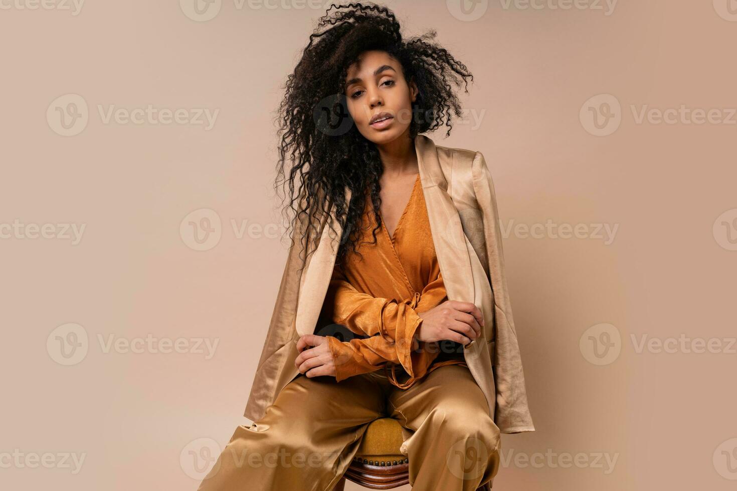 Beautiful  african model with perfect curly hairs in elegant orange blouse and silk pants sitting on vintage chair onver beige background. photo