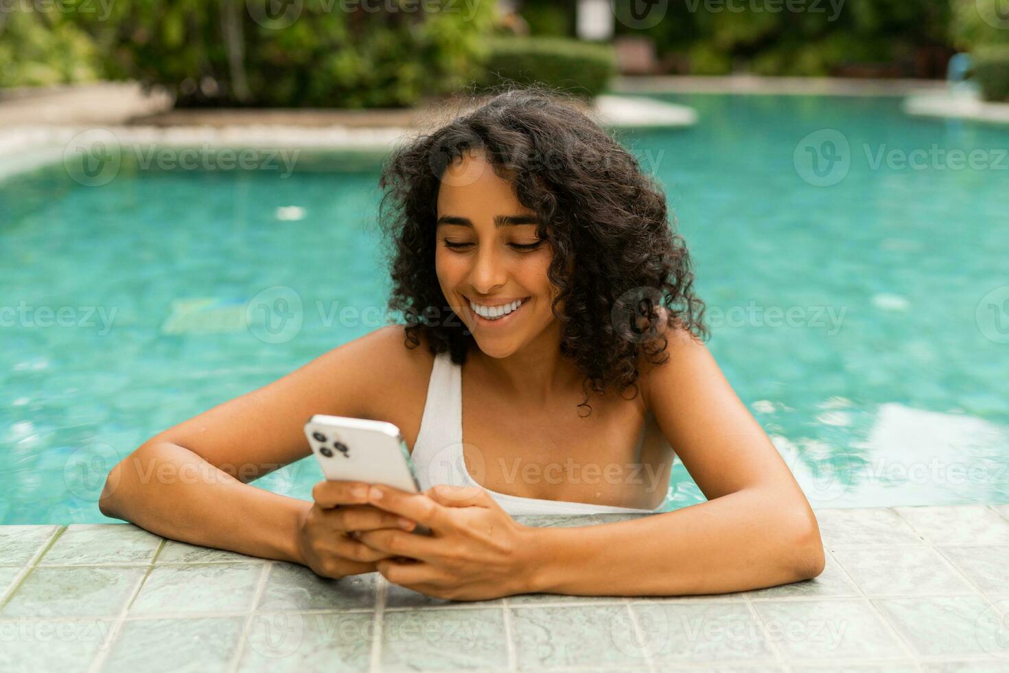 Smiling woman with curly hairstyle making self portrait by mobylephone while chilling in swimming pool. photo