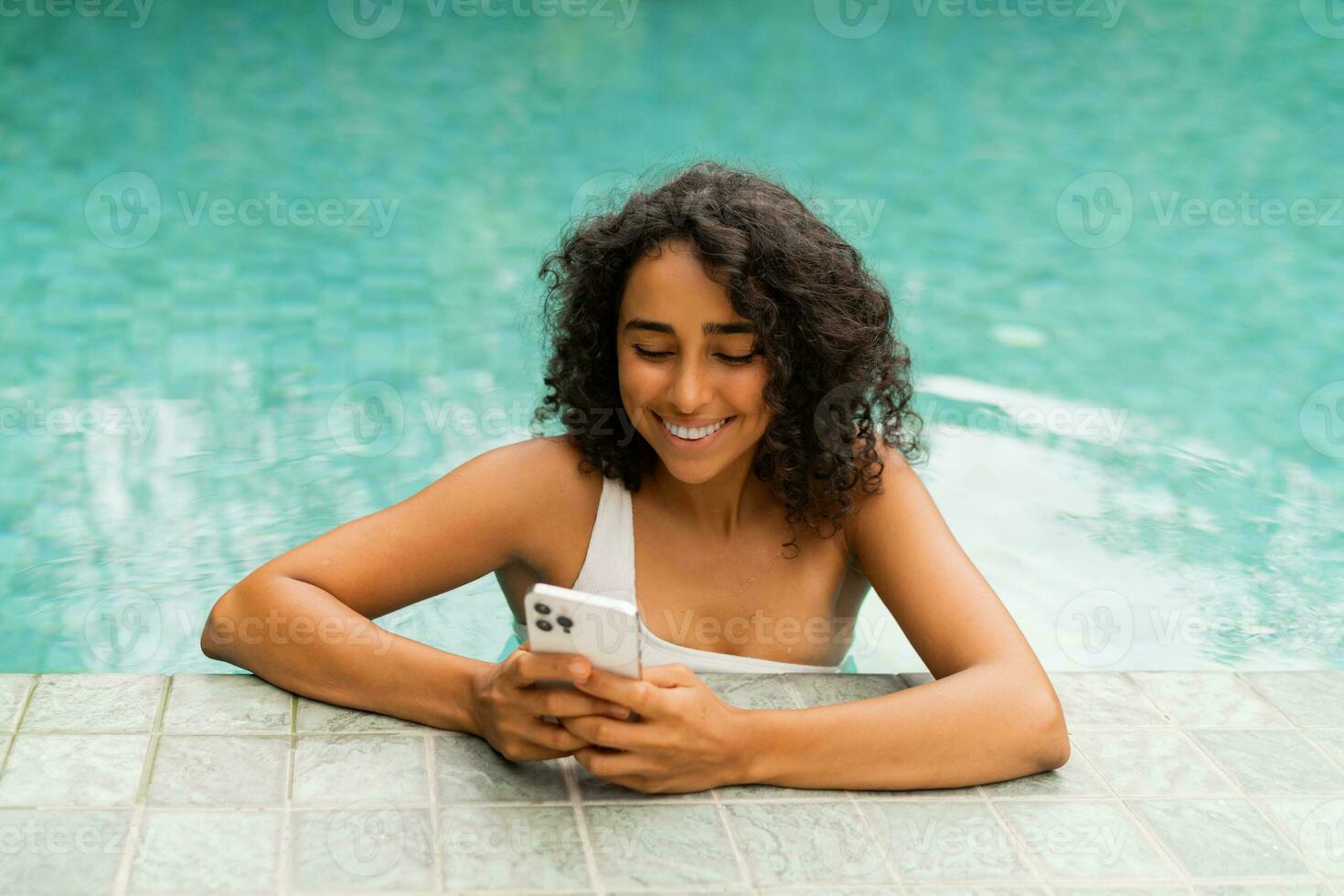 Smiling woman with curly hairstyle making self portrait by mobylephone while chilling in swimming pool. photo