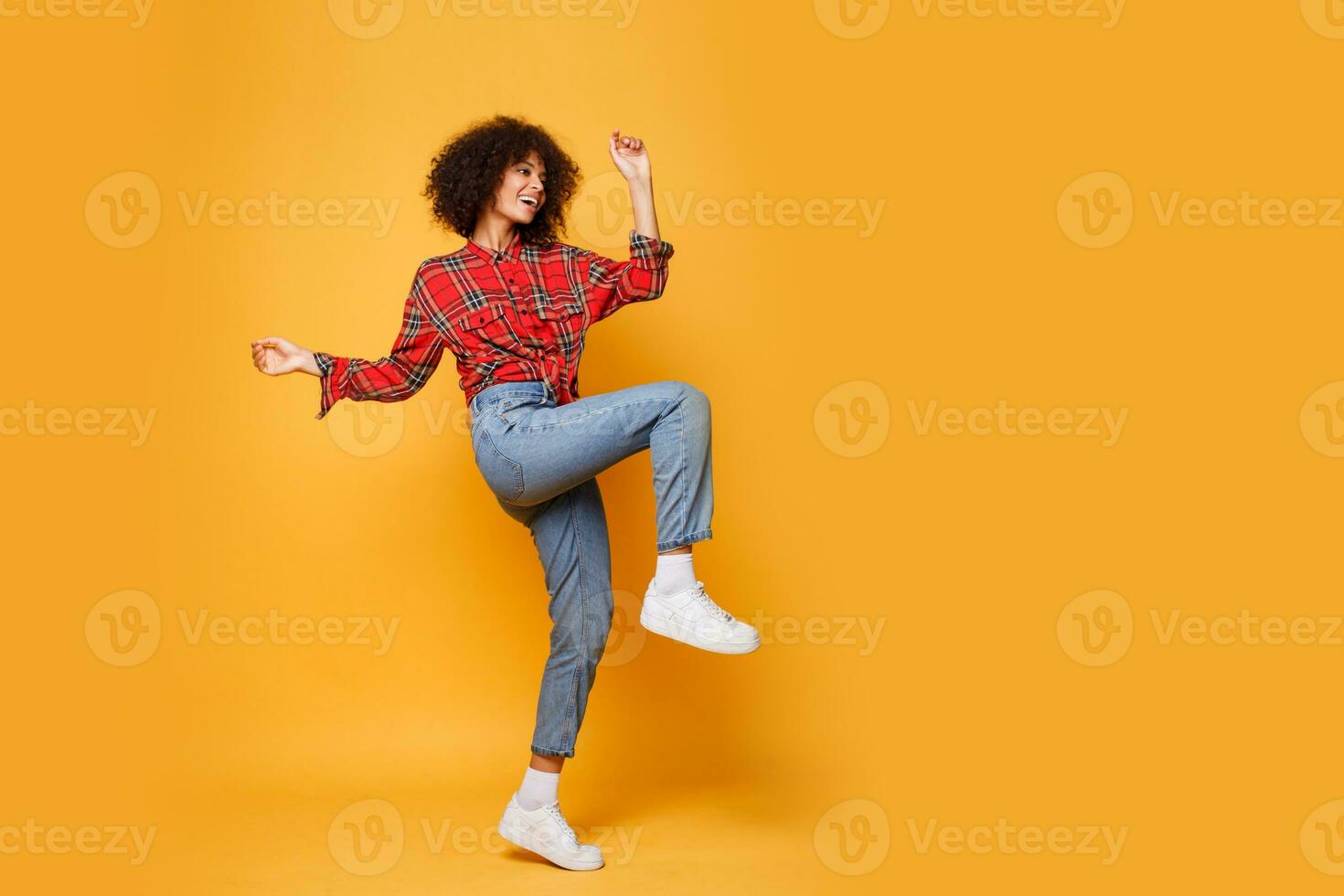 Active  girl with stylish Afro hairs  dancing on bright orange background. Studio photo of happy curly lady in  red shirt jumping and  having fun indoor. Full length.