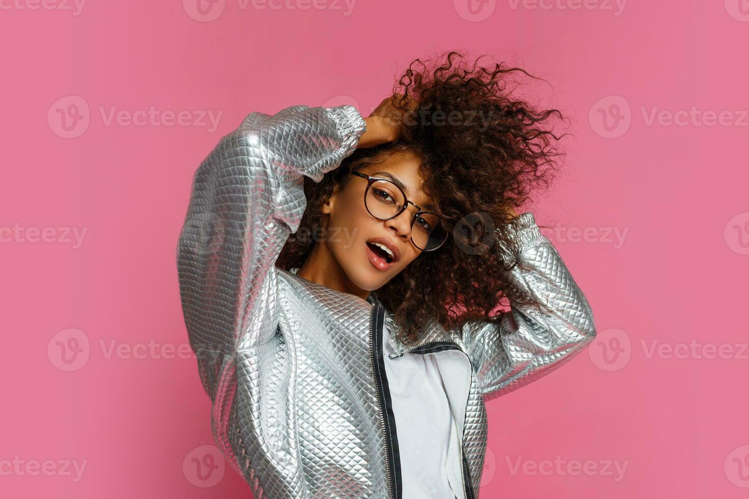 Fashionable black woman with curly hairstyle in hat and grey jacket posing on pink background in studio . photo