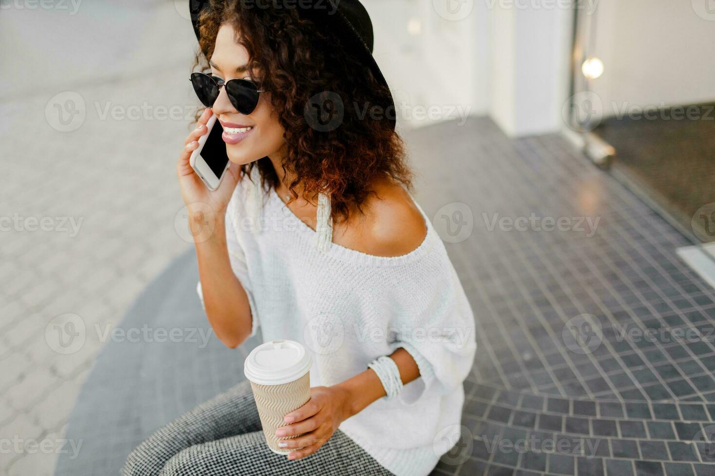 Successful black woman, blogger or store manager  talking by mobile phone during coffee break. Sitting on stairs and holding paper cup of hot beverage. White and black  fashion outfit. photo