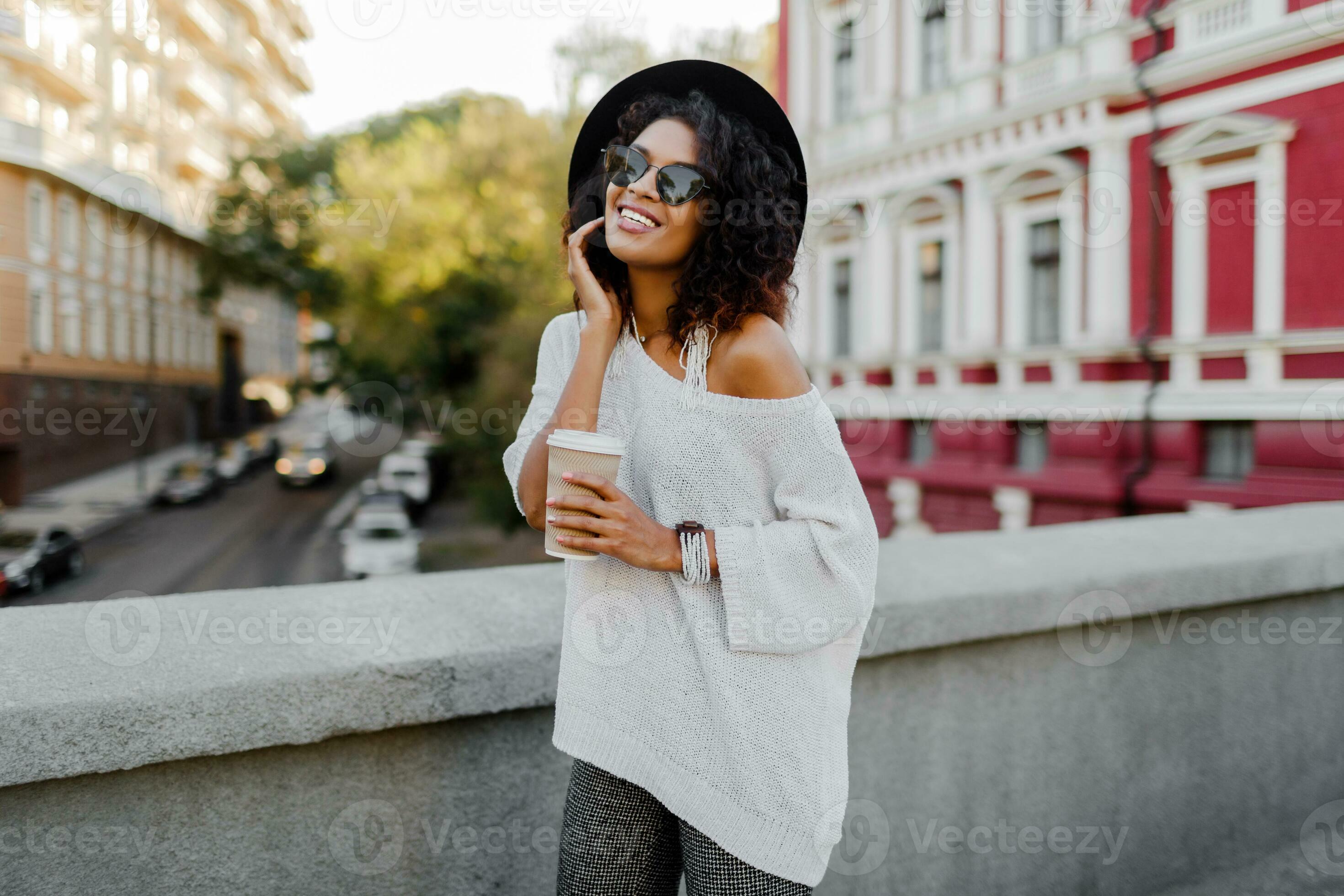 Soft toned lifestyle outdoor image of blissful black woman walking in  spring city with cup of cappuccino or hot tea. Hipster outfit. Oversize  white sweater, black hat, stylish accessories. 31416289 Stock Photo