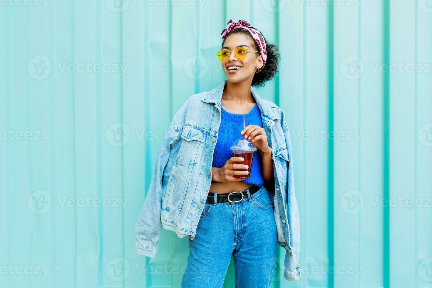 Happy mixed race female with stylish Afro hairs posing outdoor , holding cherry lemonade , wearing jeans jacket and blue wool sweater. photo
