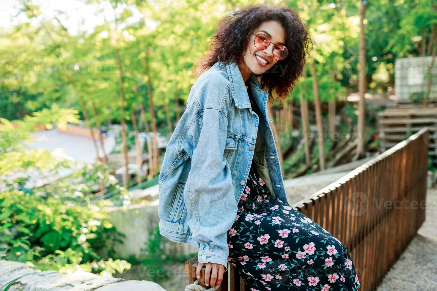 sonriente mujer con afro peinado sentado en un cerca en verano soleado parque . vistiendo pantalones chaqueta, vestir con floral impresión y zapatillas . foto