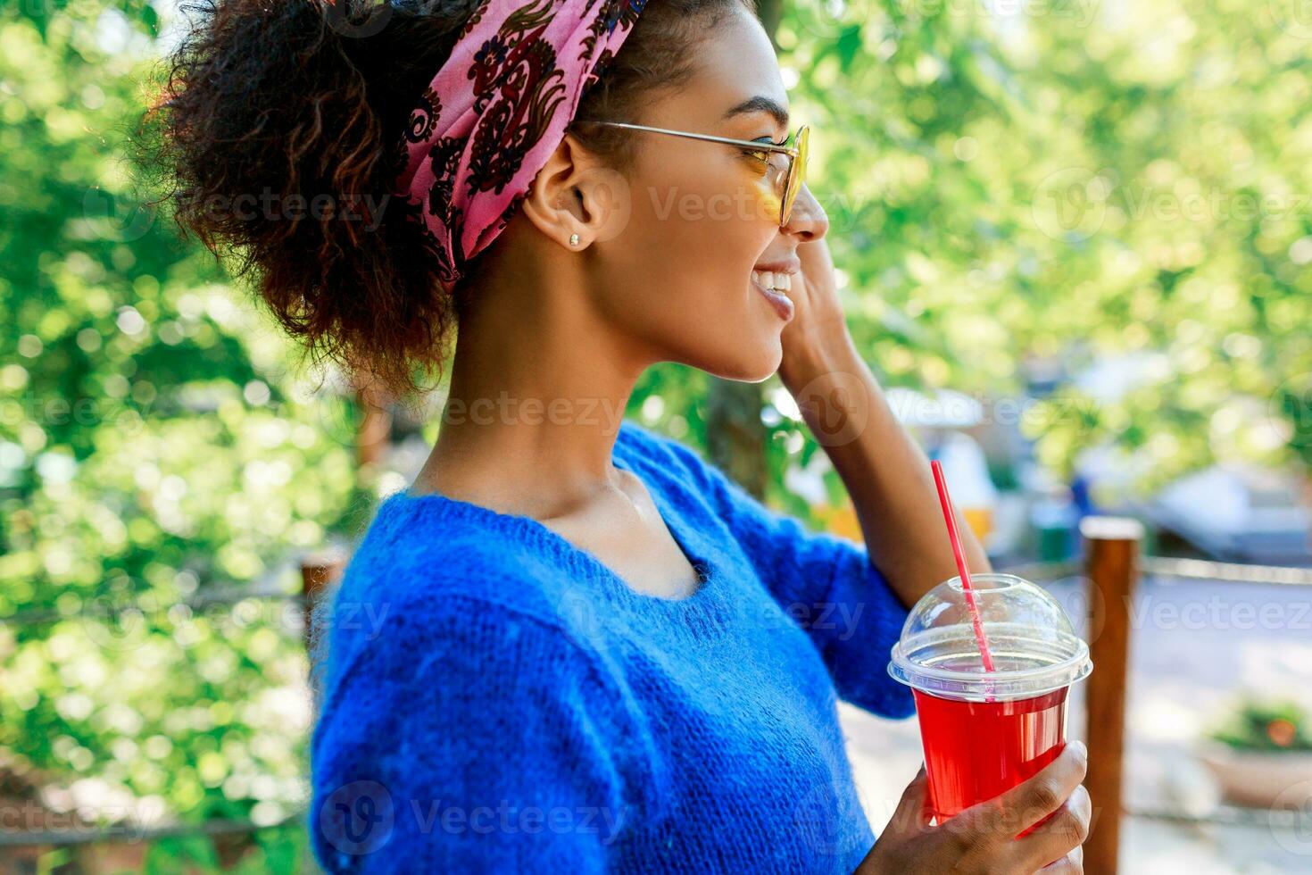 Profile of  happy  black  female  with stylish headband looking on horizon  and  holding cherry lemonade on hands. Summer park background. photo