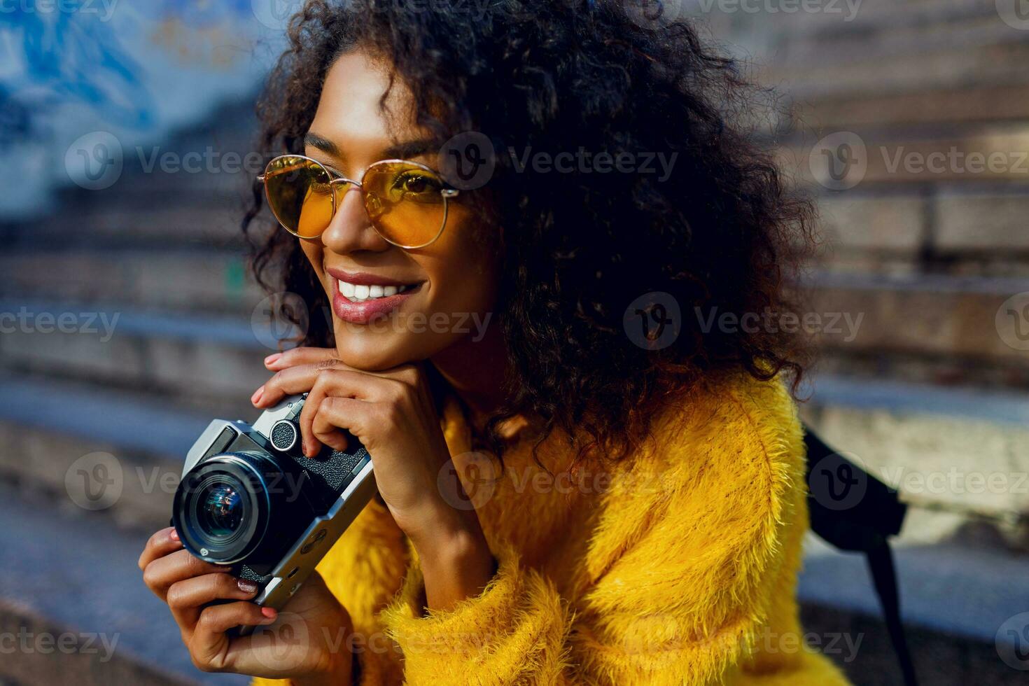 Stylish inspired African woman sitting on stairs and dreaming. Holding retro film camera. Urban background. photo