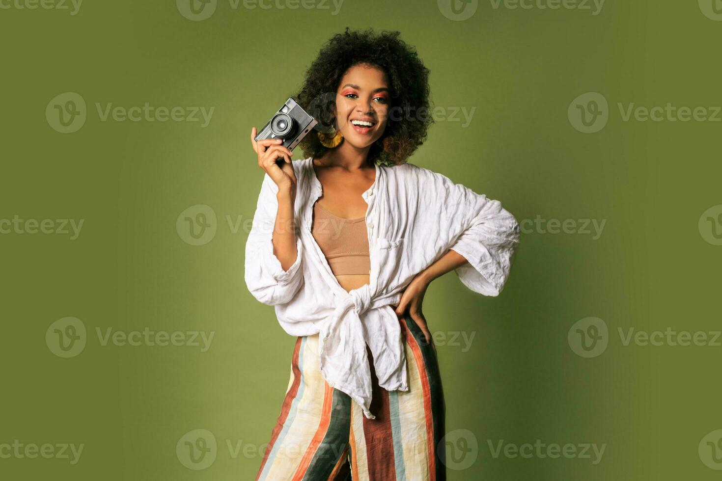 Afican woman holding retro photocamera and  laughing over green background in studio. photo