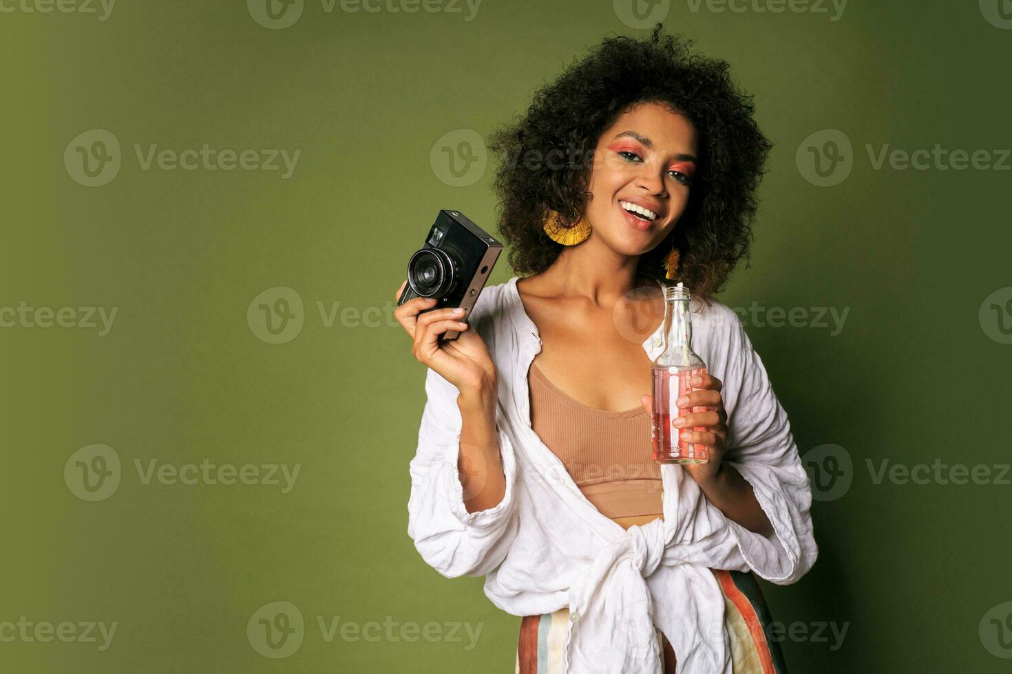 Pretty african woman with stislish hairstyle posing in studio, drinking lemonade from straw. Summer style. Green background. Bright make up. photo