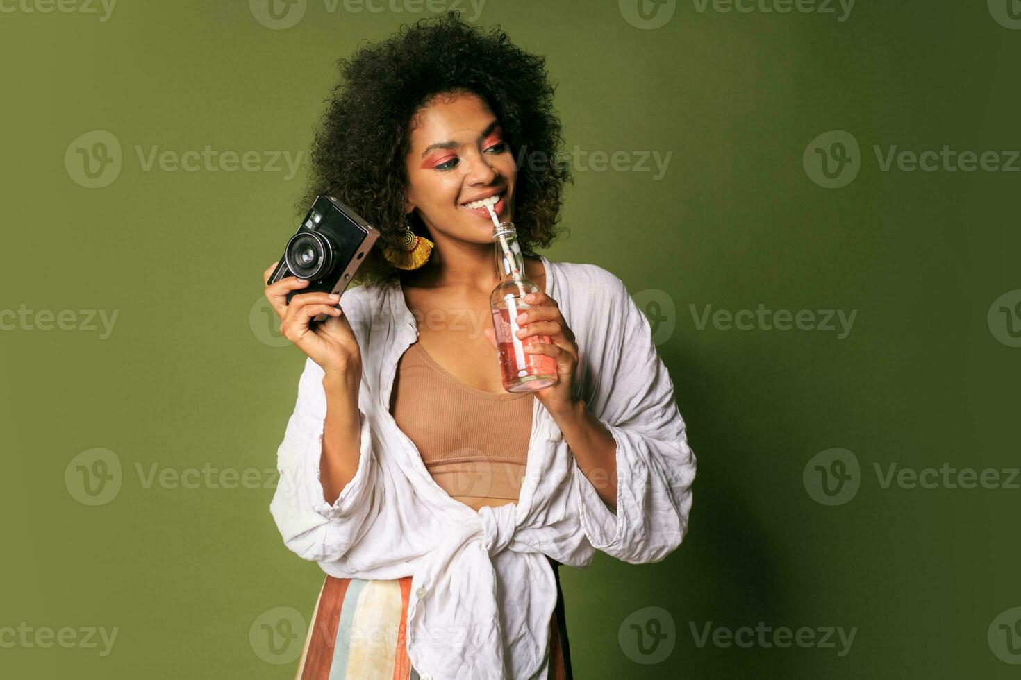 Pretty african woman with stislish hairstyle posing in studio, drinking lemonade from straw. Summer style. Green background. Bright make up. photo