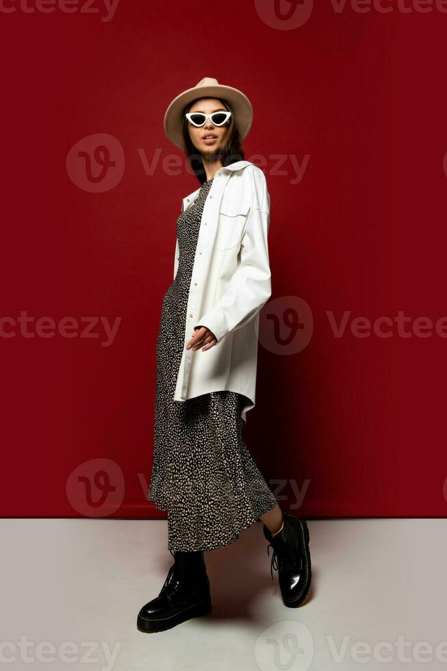 Elegant feshionable woman in white ovesize jacket and dress posing in studio on red background. Ankle boot in black leather. Full lenght. photo