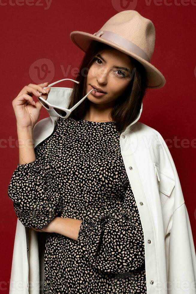 Fashion portrait of elegant   brunette woman with  dressed  white  coat  and beige hat holding sunglasses.  Autumn or winter trendy fashion style.  Red background. photo