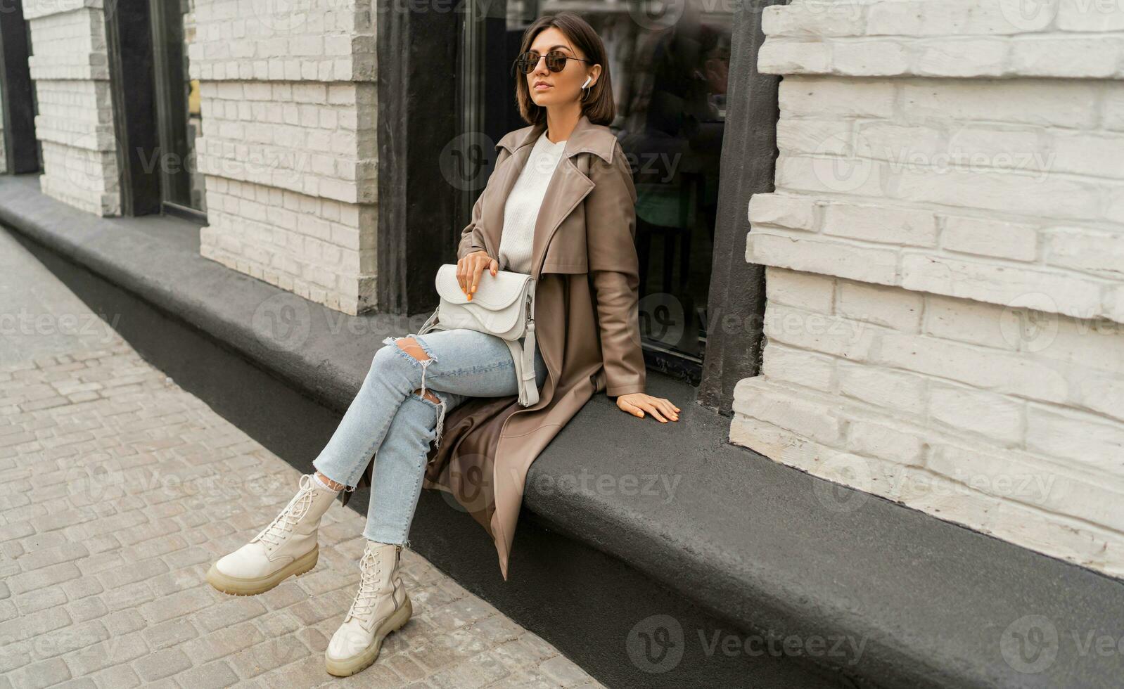 elegante de moda mujer con corto bruneete pelos vistiendo de moda Gafas de sol, beige color abrigo, blanco texturizado cuero hombro bolsa, caminando en calle de europeo ciudad. foto