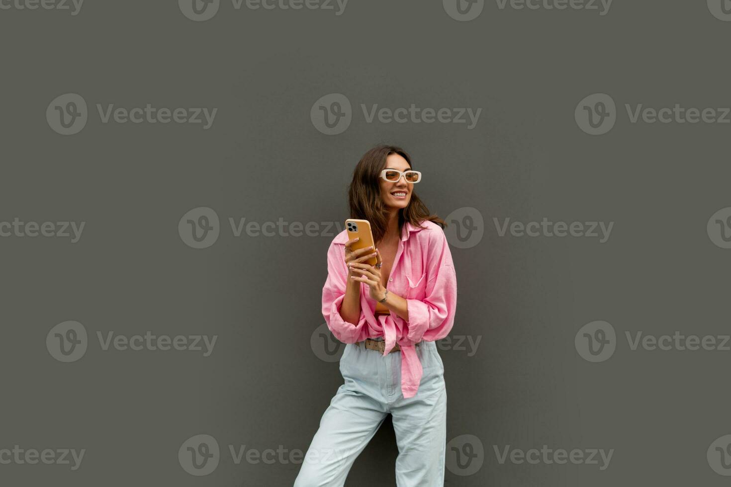 Attrcative playful woman in trendy sunglasses and casual  clothes posing over grey background. Using smartphone. photo