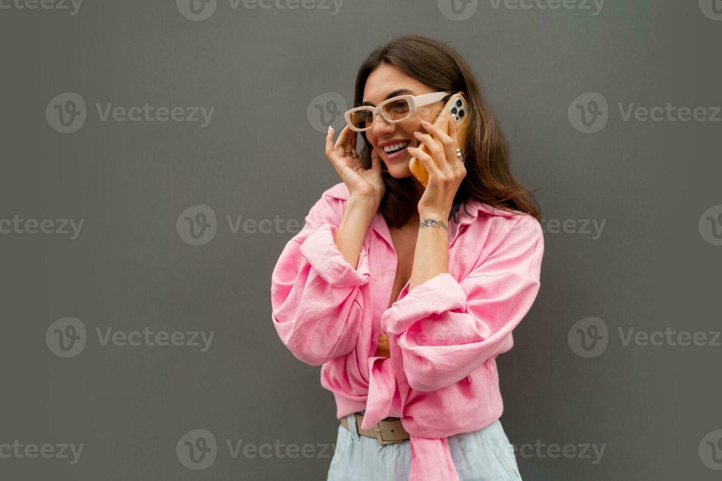 atractivo juguetón mujer en de moda Gafas de sol y casual ropa posando terminado gris antecedentes. bonito niña hablando por móvil teléfono. foto