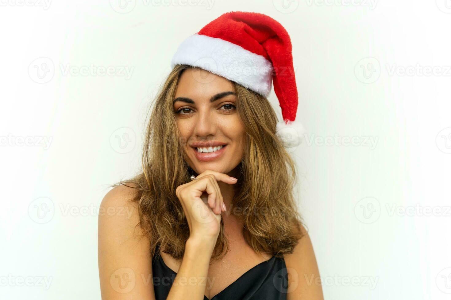Close up portrait of beautiful brunette smiling  woman in  red santa hat posing on white background. Wearing elegant black dress. photo