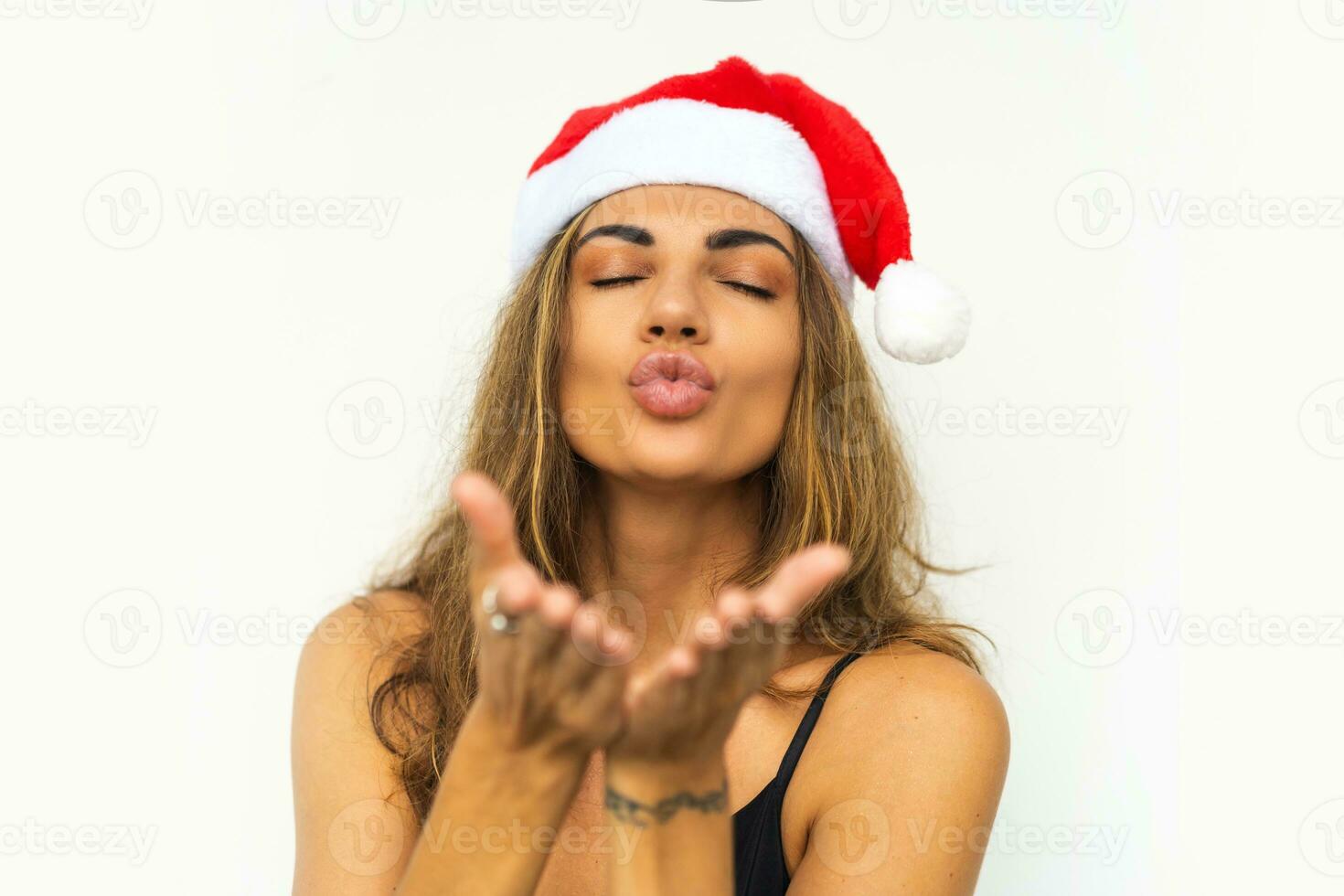 bonito mujer con en Papa Noel sombrero y elegante negro vestir enviar Beso a cámara y posando en blanco antecedentes en estudio. foto