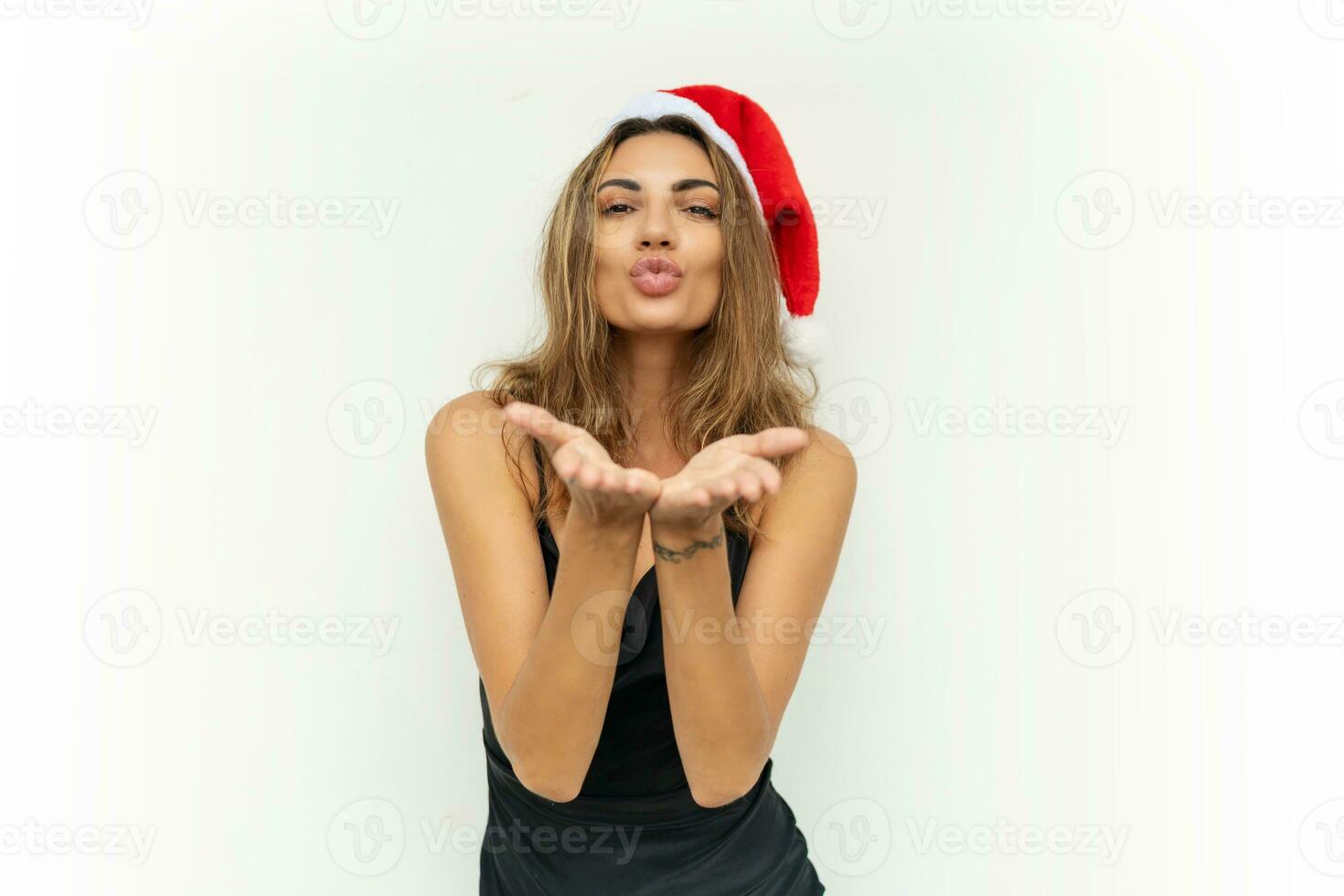 Happy woman in santa hat and sexy black party dress send kiss to camera. Posing on white background. photo