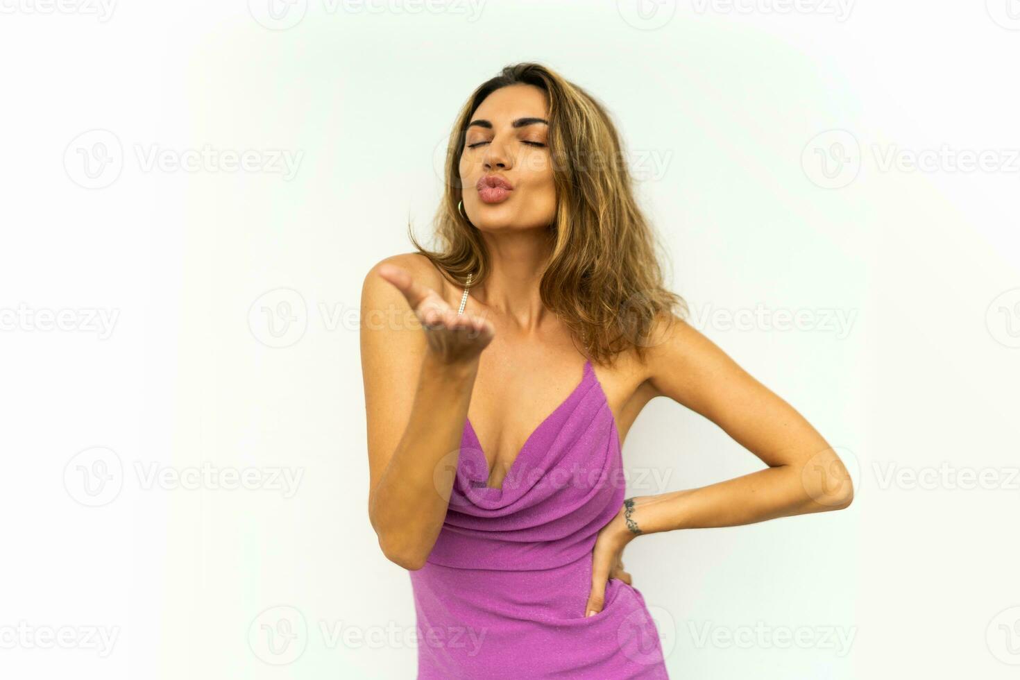 elegante europeo mujer enviar Beso a cámara vistiendo rosado brillante vestir y ondulado pelos posando en blanco antecedentes. foto