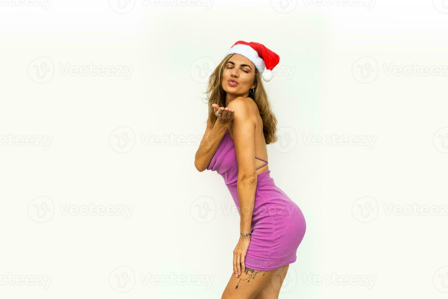 bonito mujer en rojo Papa Noel sombrero enviar Beso a cámara vistiendo rosado brillante vestir . ondulado pelos posando en blanco antecedentes. foto