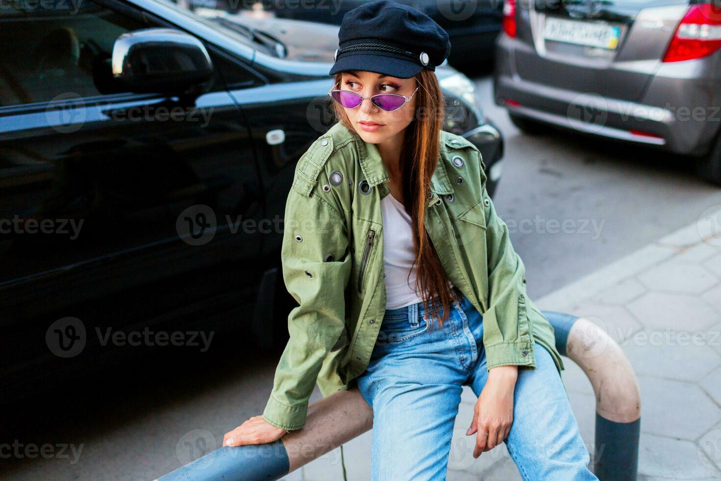 Fashionable woman with long brunet hairs posing outdoors in big  city near road . Wearing stylish green jacket , black cap and purple sunglasses. photo