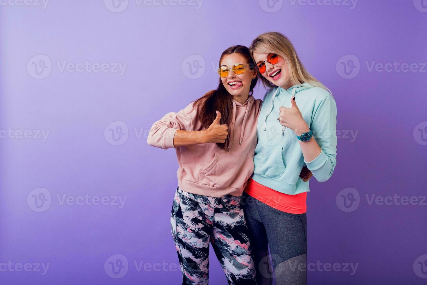 Two girls with surprise face  staying  over purple wall. Wearing fashionable hoodies and cool glasses. photo