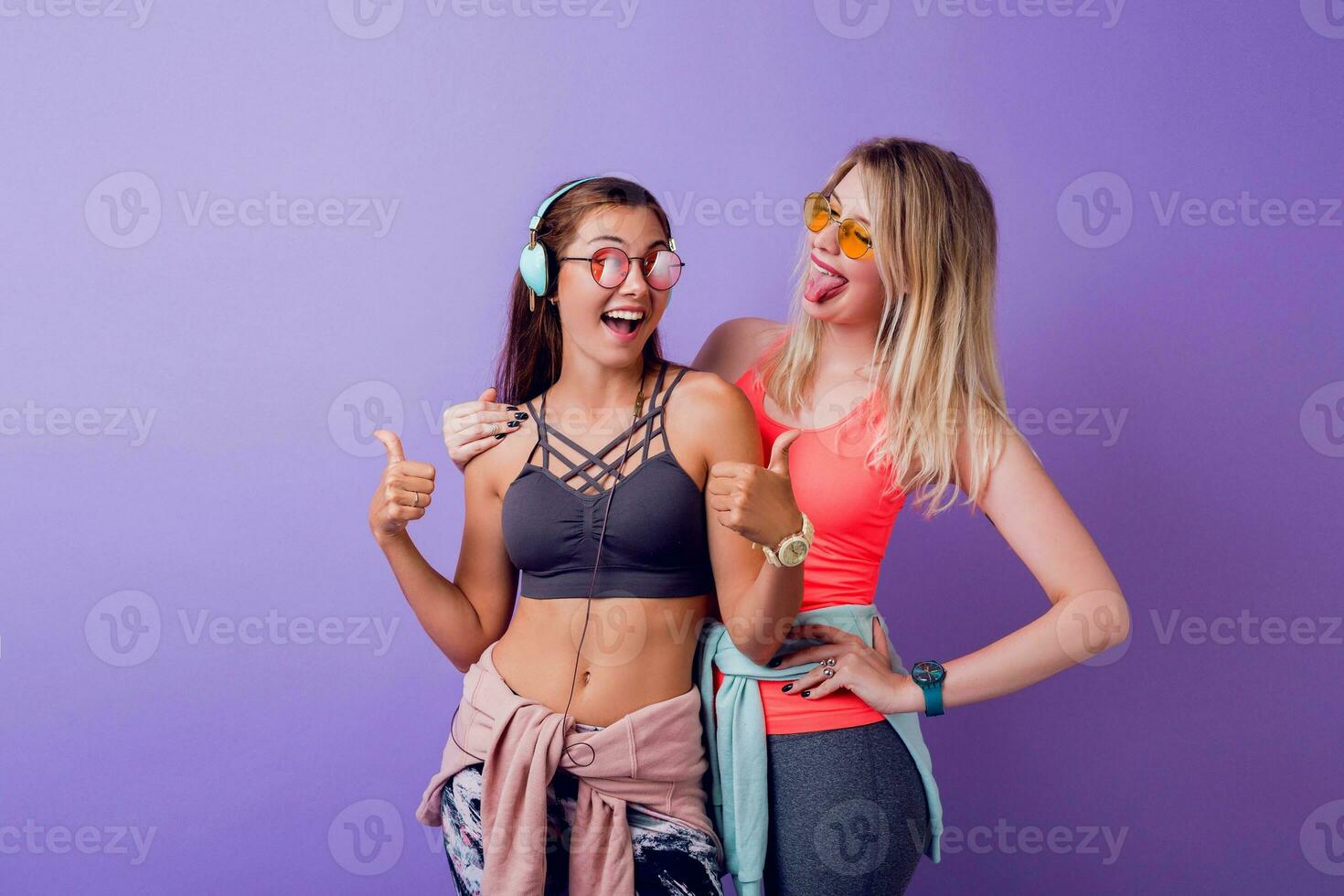 dos bonito muchachas en de moda ropa deportiva posando en púrpura antecedentes en estudio. morena y rubio mujer en pie juntos. aptitud y activo Moda concepto. foto