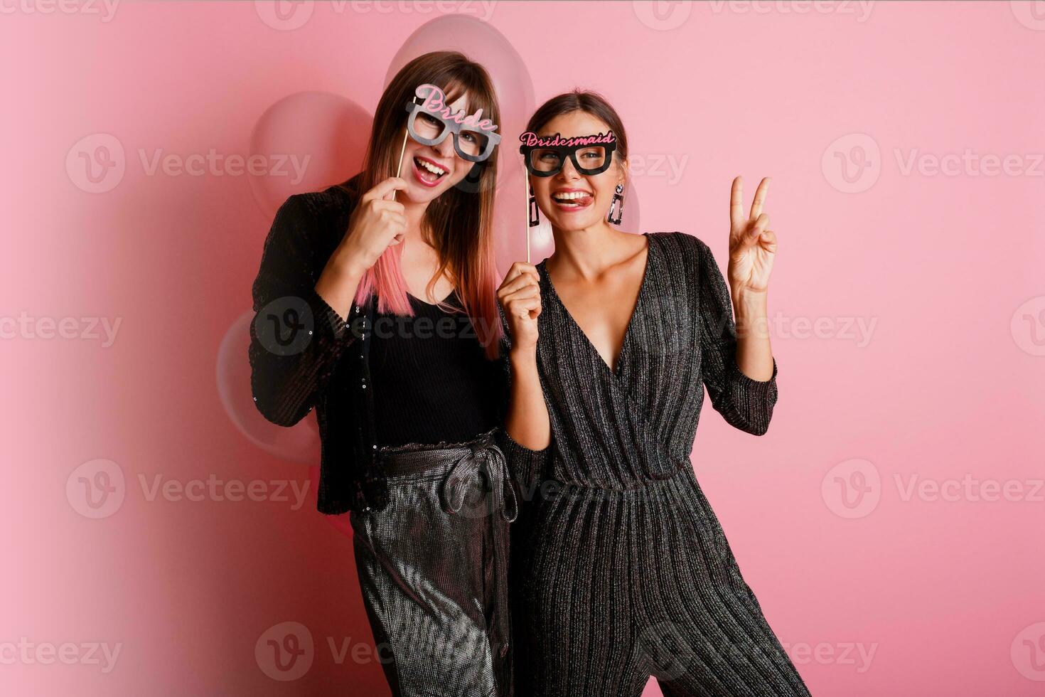 Two women , best friends  celebrating hen party, posing with bridal photo props on pink background.