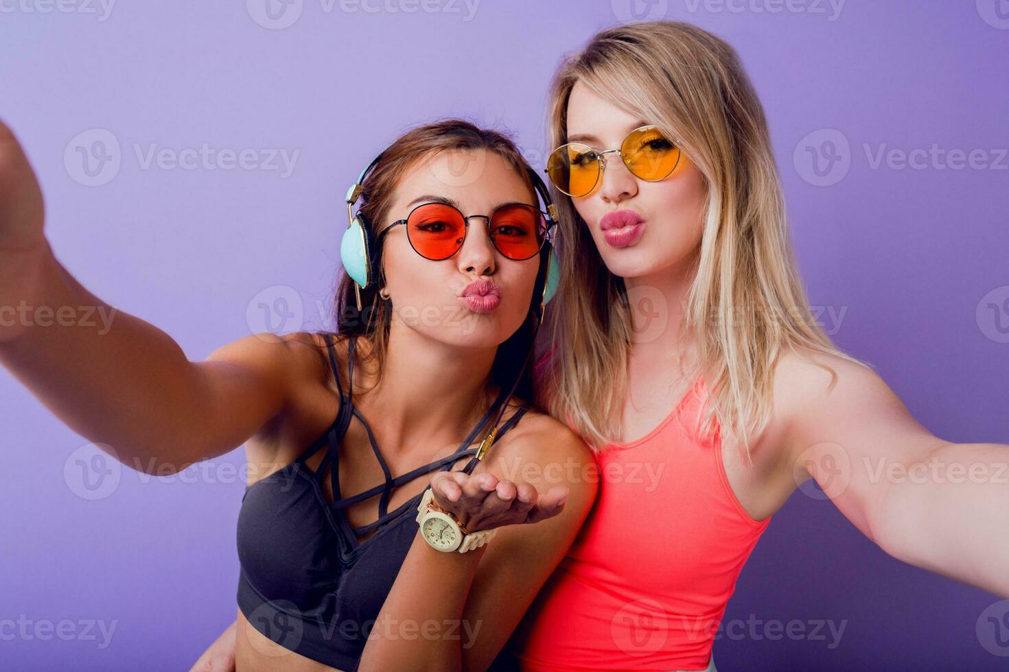 Fitness girls send air kiss and taking self portrait by mobile phon.  Wearing bright pink activewear. Stylish sunglasses. Posing on purple background. photo