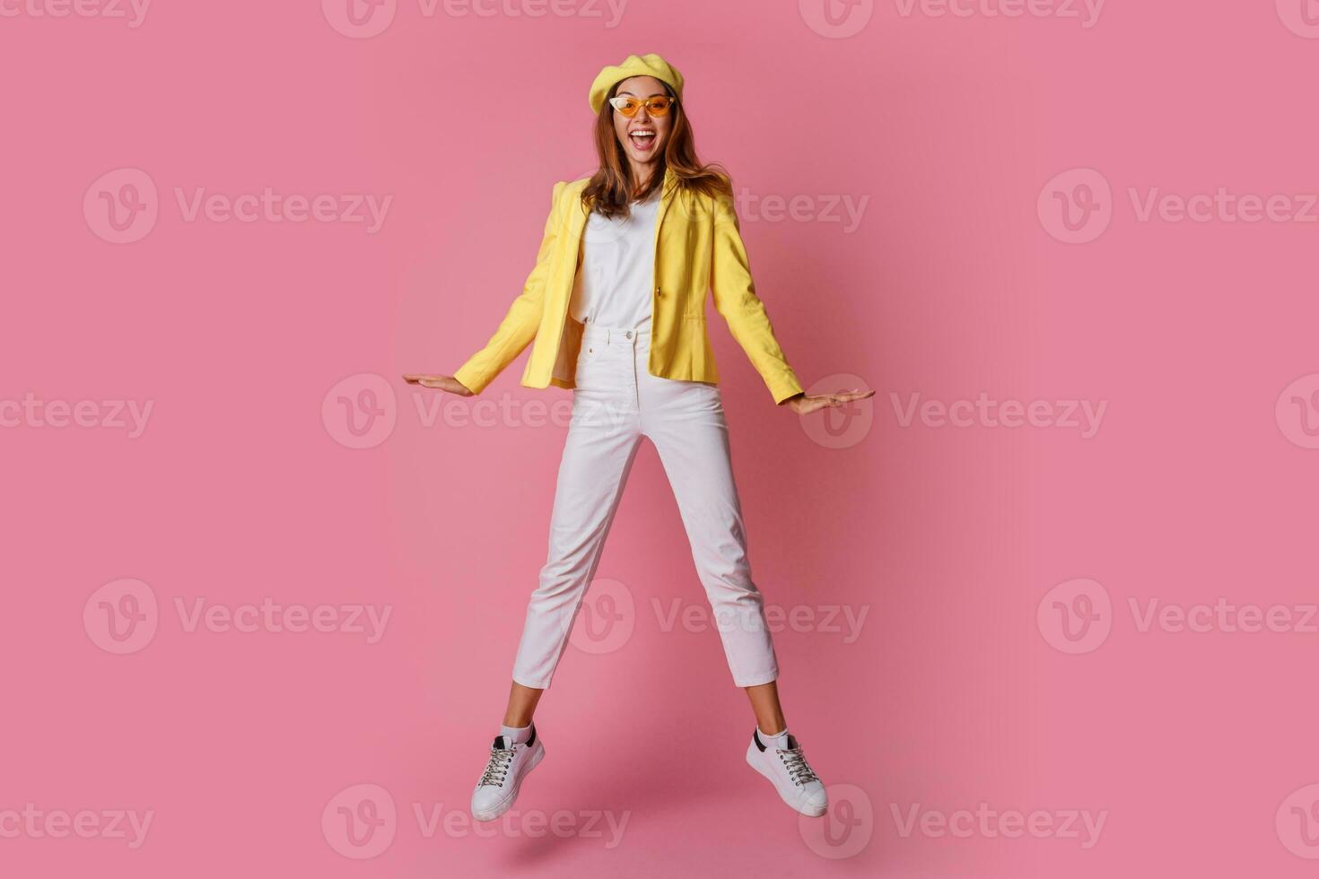 Inspired brunette girl  in yellow jacket and beret jumping in studio on pink background. Wearing white sneakers and jeans. Happiness mood. Fashion trends. photo