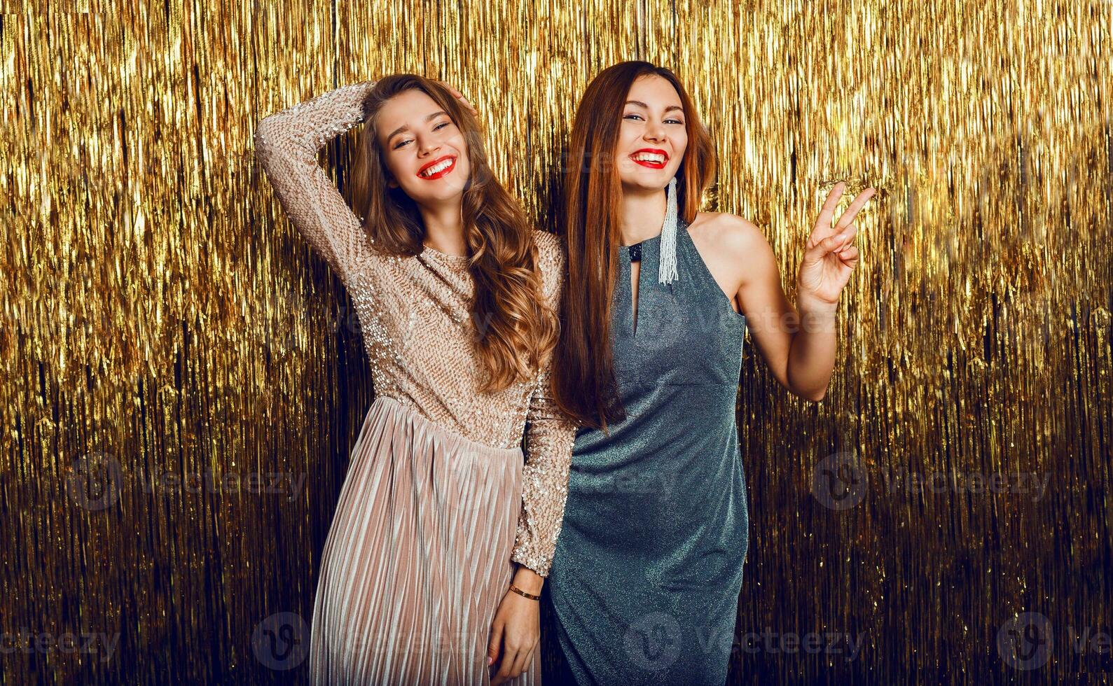 Two celebrating girls in elegant evening dress posing on golden  sequence  background. Having fun.  Bright make up, red lips. New year party mood. photo