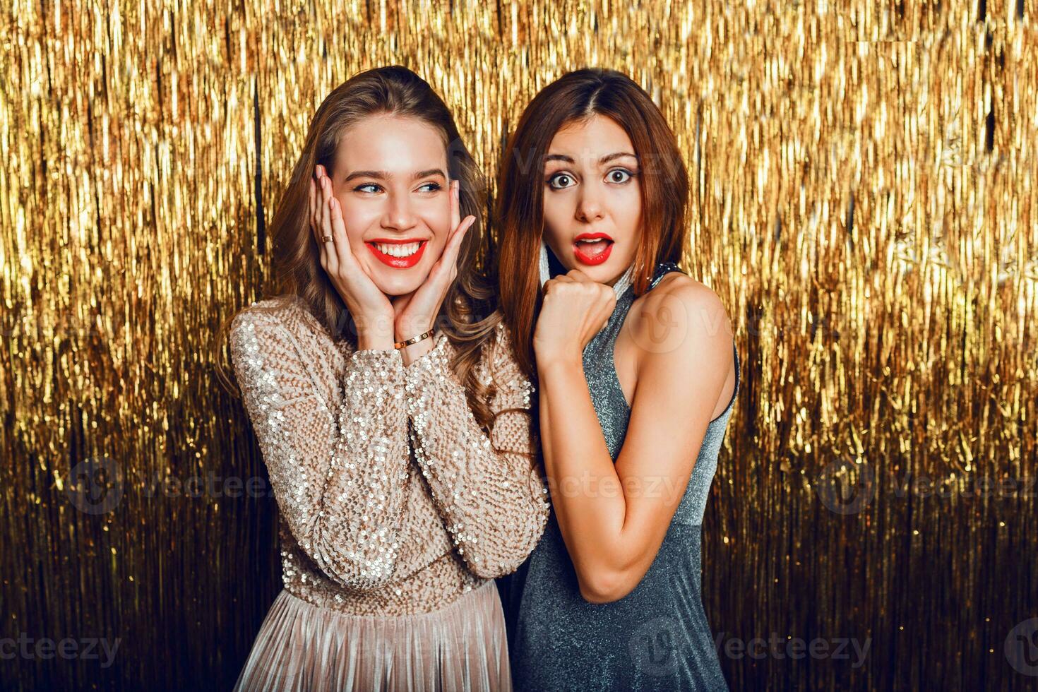 cerca arriba estudio imagen de dos increíble sexy celebrando muchachas con rojo labios, sorpresa rostro, riendo , posando en dorado brillante antecedentes. nuevo año fiesta humor. foto