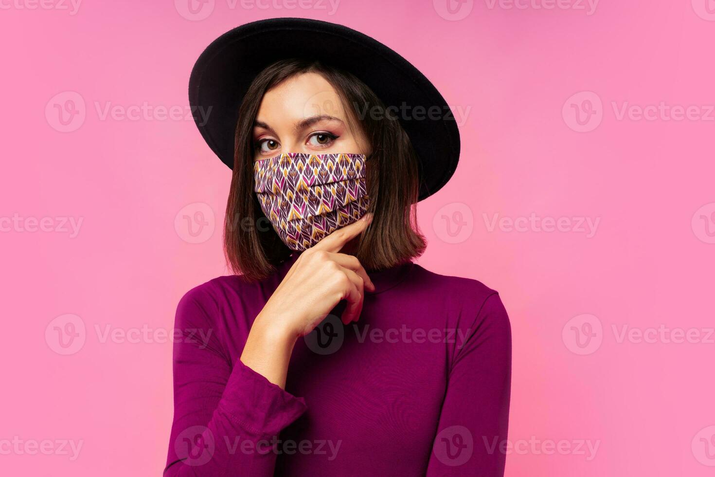 Beautiful woman wearing stylish protective face mask. Pink background. Black hat. Studio photo. photo