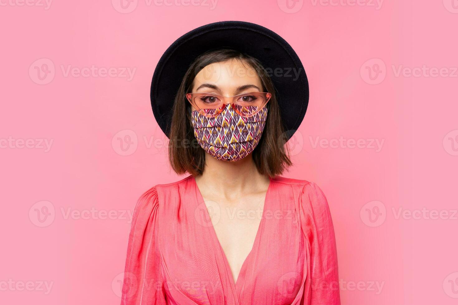 European model dressed protective stylish face mask. Wearing black hat and sunglasses. Posing over pink background in studio. photo