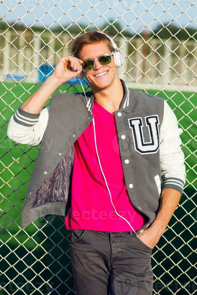 Outdoor fashion portrait of handsome guy in stylish  spring sportive outfit, listening favorite music  and posing near sports field. Bright contrast colors. photo