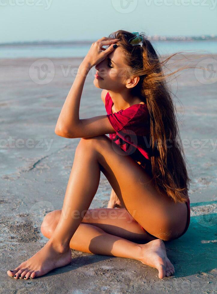 Tanned slim fitness   lady  thoughtfully sitting on the beach  in a red T-shirt  and bikini . Have a sunbath  evening summer sun. photo