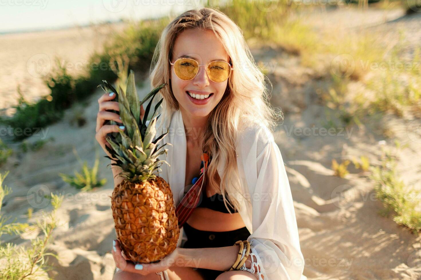 Picture of laughing pretty woman in trendy summer outfit with pineapple relaxing on sunny beach. photo