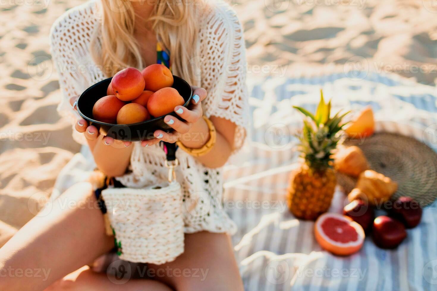 Beautiful blond girl holding plate with peach and relaxing on the evening tropical beach, sunset colors. Sitting on towel. Tropical fruits. Wearing stylish headband and boho dress. Healthy life. photo