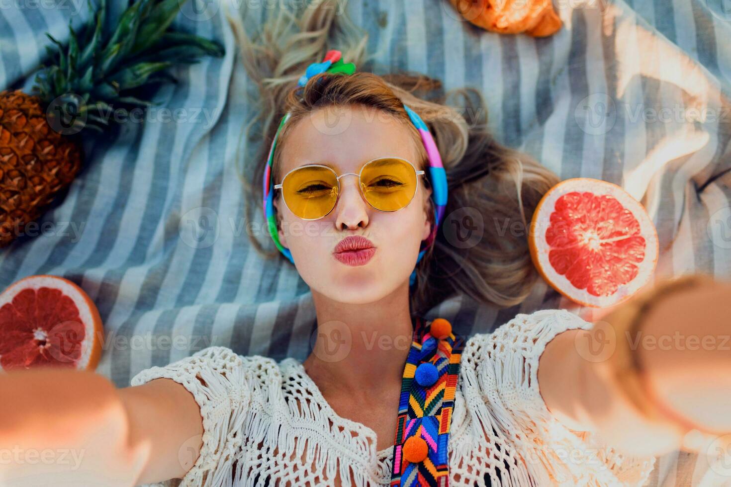 Excited girl making selfie on beach towel. Boho accessories. Yellow sunglasses.Top view. photo