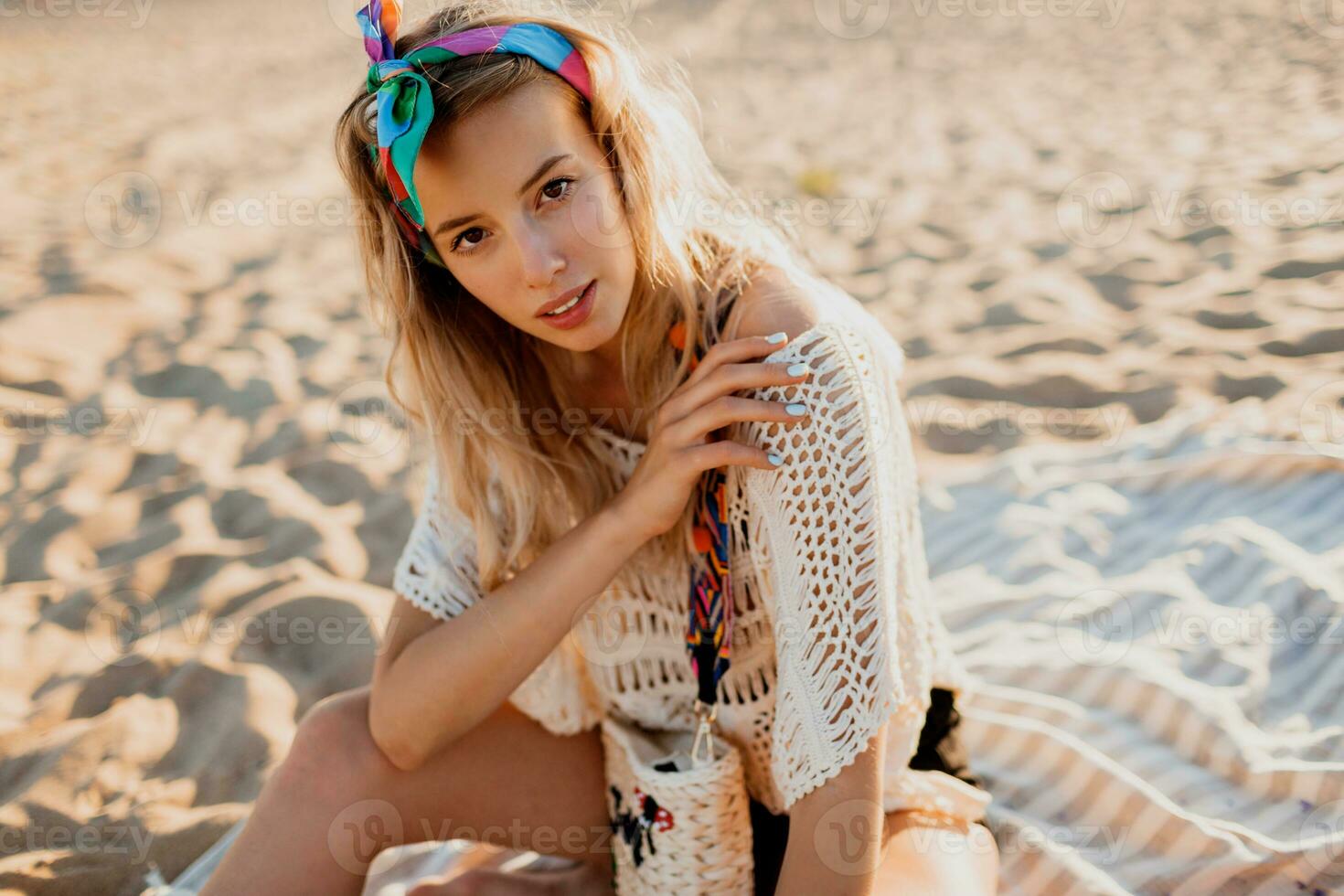 Warm summer portrait of stunning graceful woman enjoying sunset near ocean, sitting on sand. Perfect smile, wearing white boho dress and colorful headband. photo